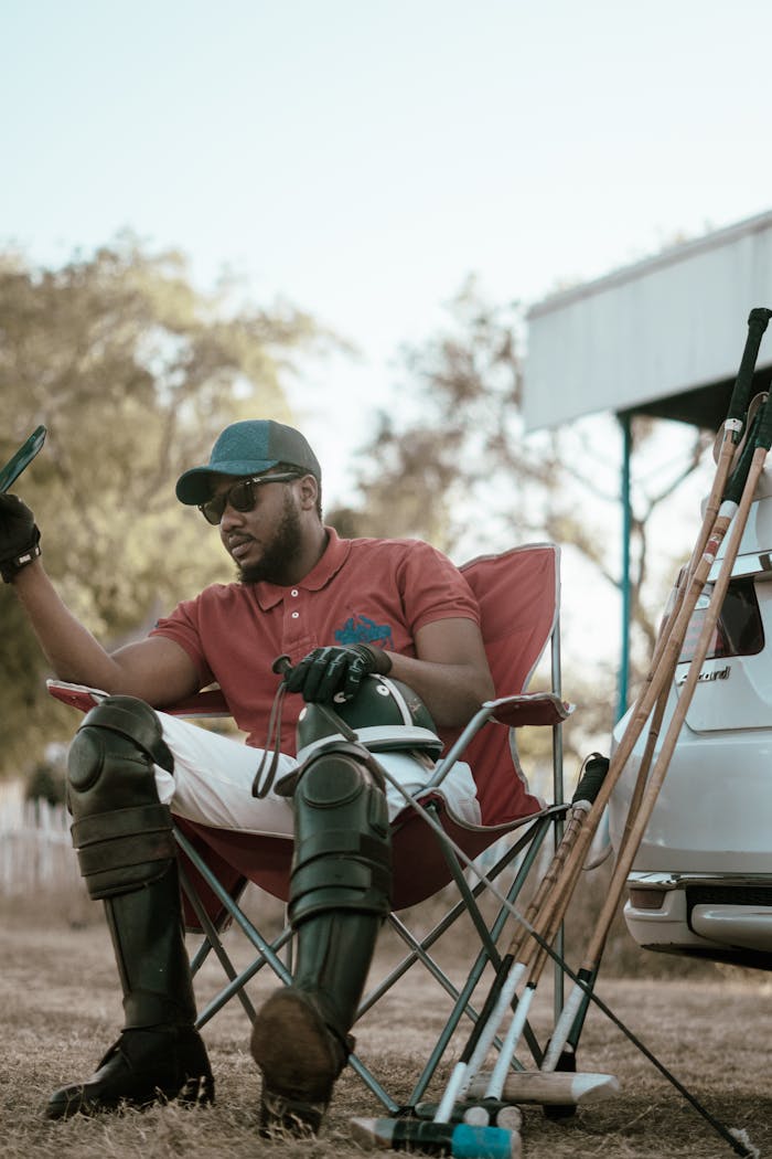 Man in polo gear seated in a chair outdoors, sporting sunglasses and cap near polo sticks.