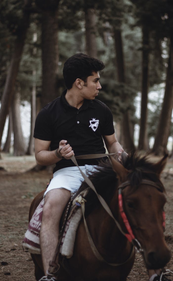 A young man in a black polo shirt riding a brown horse through a serene forest setting.